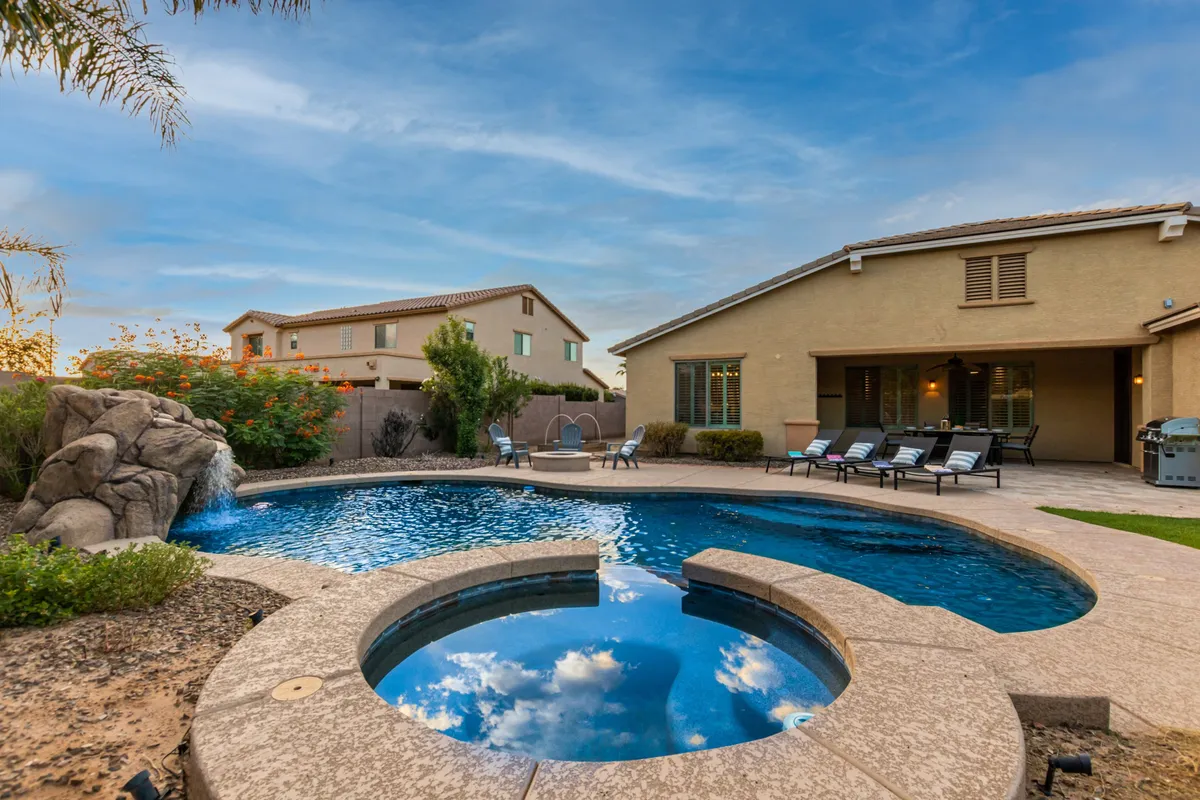This image showcases the backyard's sparkling swimming pool surrounded by lounge chairs, a cascading waterfall feature, and an inviting outdoor dining area, all set against a tranquil evening sky. The setup visually captures a blend of leisure and gathering spaces for both family fun and relaxation.