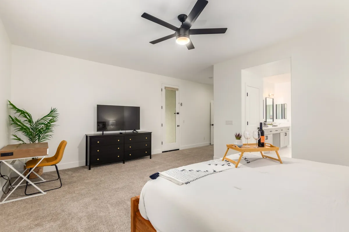 A spacious bedroom is shown with a bed adorned in a decorative throw, a TV on a modern dresser, a small desk with a chair, an indoor plant, and a doorway leading to an en-suite bathroom. Natural light fills the room, and the decor is stylish yet soothing.