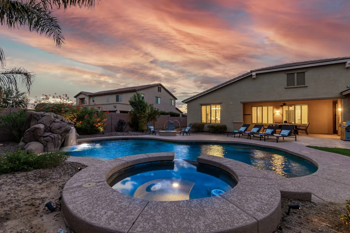 A peaceful backyard scene featuring a swimming pool, spa, lush landscaping, and a vivid sunset sky. There are spaces for relaxation, gatherings, and unwinding after a day of activity.