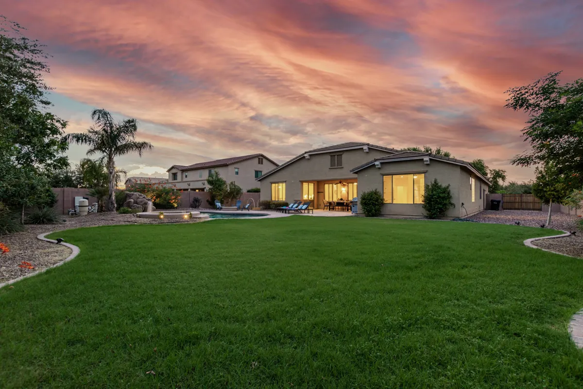 A vibrant backyard features a lush green lawn that leads to a spacious swimming pool. The pool is framed by comfortable lounge chairs and landscaping, while the house's exterior and a glowing sunset sky complete the inviting setting—an ideal space for outdoor gatherings and family barbecues.