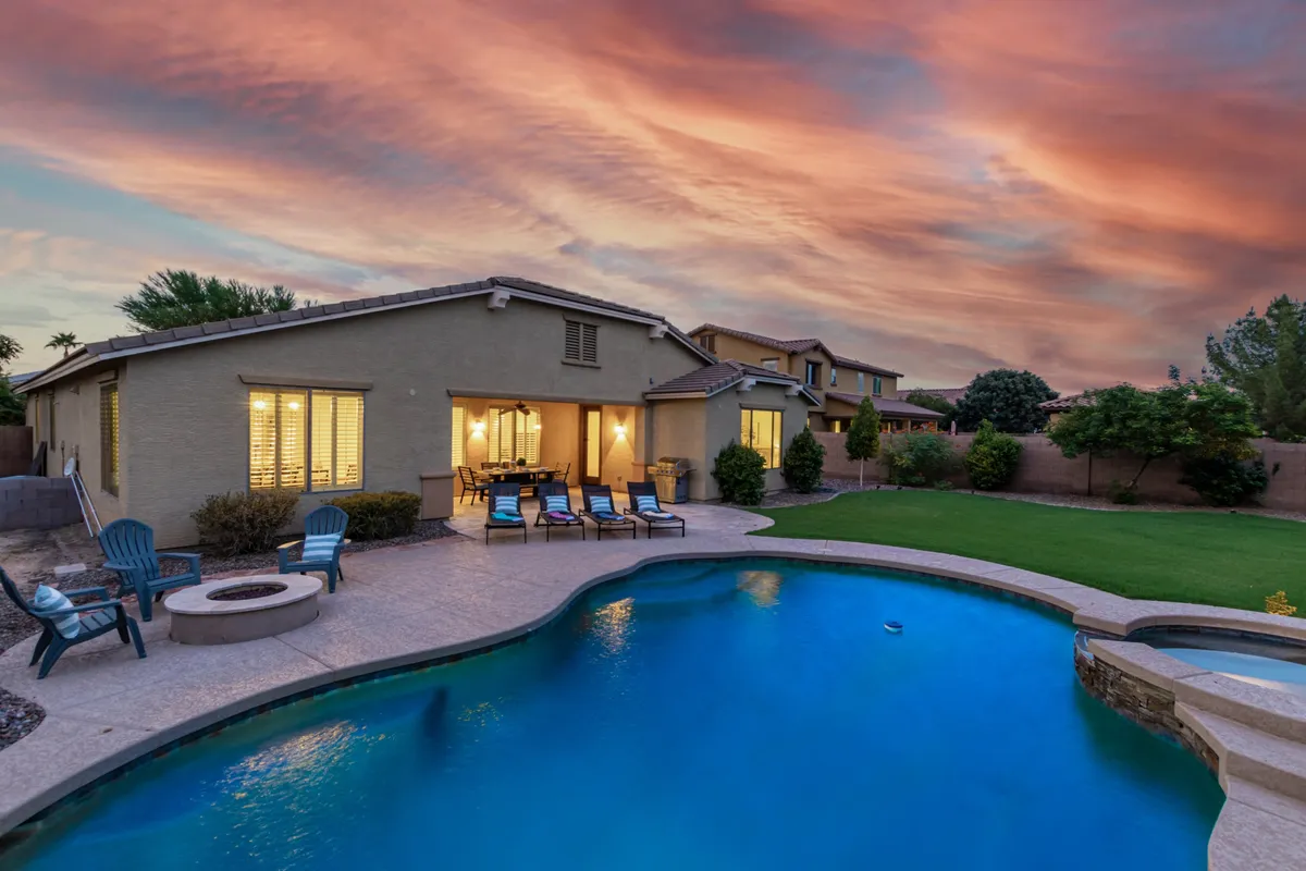 The scene shows a serene backyard with a sparkling swimming pool, an outdoor seating area, and a glowing fire pit, all illuminated by the backdrop of a gorgeous sunset sky. Lounge chairs and well-manicured landscaping add to the resort-like vibe. The setting is designed for relaxation, evening gatherings, outdoor dining, and stargazing in a tranquil, private environment.