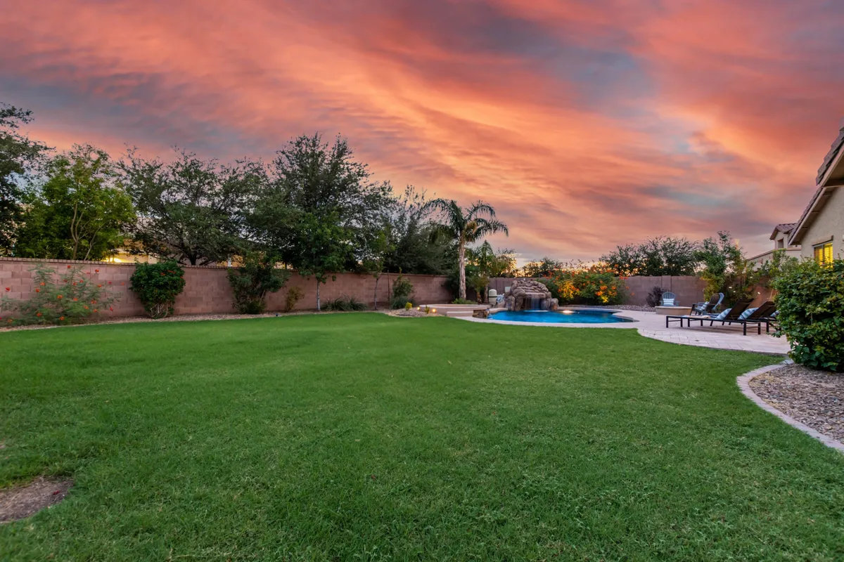 A lush green lawn frames a shimmering pool with a sunset sky in the background, accompanied by a cozy patio seating area. The scene feels relaxed and inviting, highlighting play areas for families and comfortable lounging options.