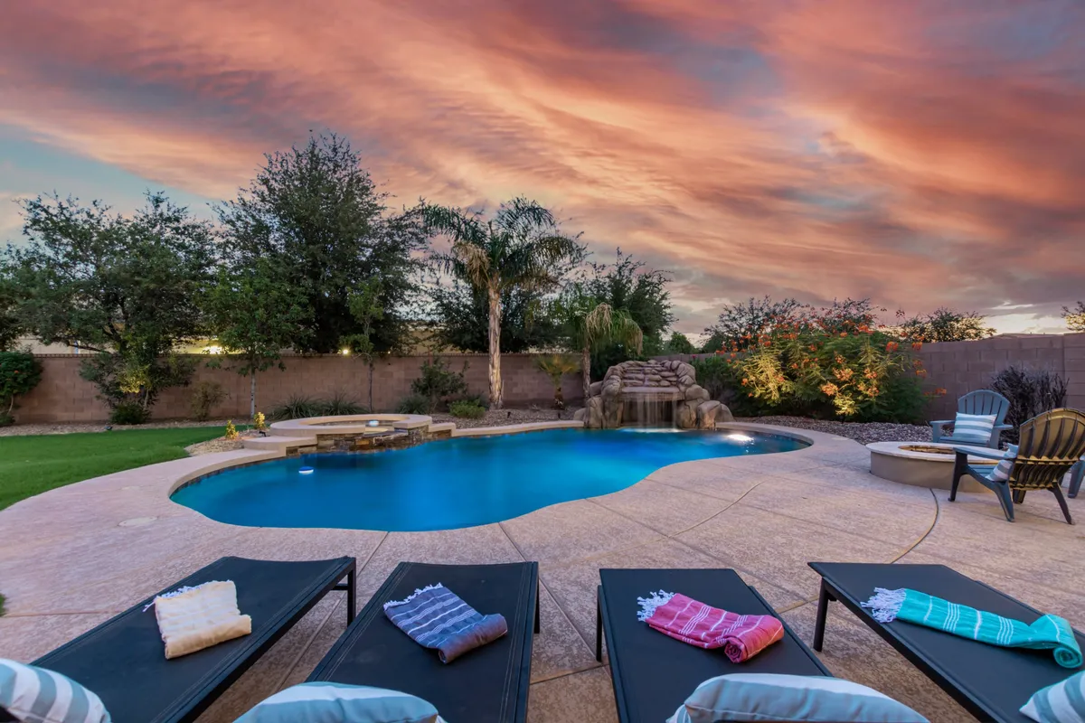 This exterior image features an inviting swimming pool with a gentle waterfall, surrounded by comfortable lounge chairs and lush landscaping, all under a vibrant sunset sky. The tranquil setting exudes resort-like luxury, perfect for swimming, sunbathing, or socializing poolside in a private, upscale environment.