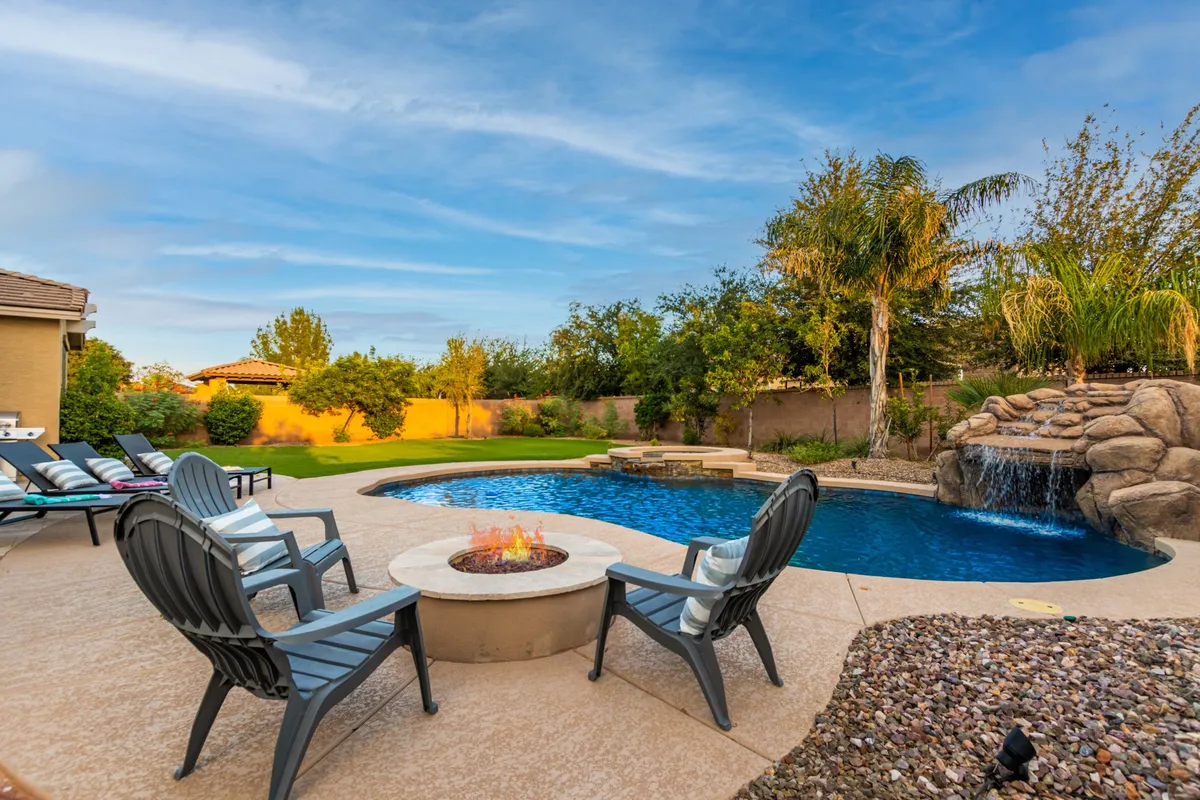 A beautifully landscaped backyard features a sparkling swimming pool with a rock waterfall, surrounded by lush greenery and lounge chairs. In the corner, a cozy fire pit area invites relaxing evenings, while the pool area is bathed in sunset light, creating a tranquil atmosphere perfect for family fun and relaxation.