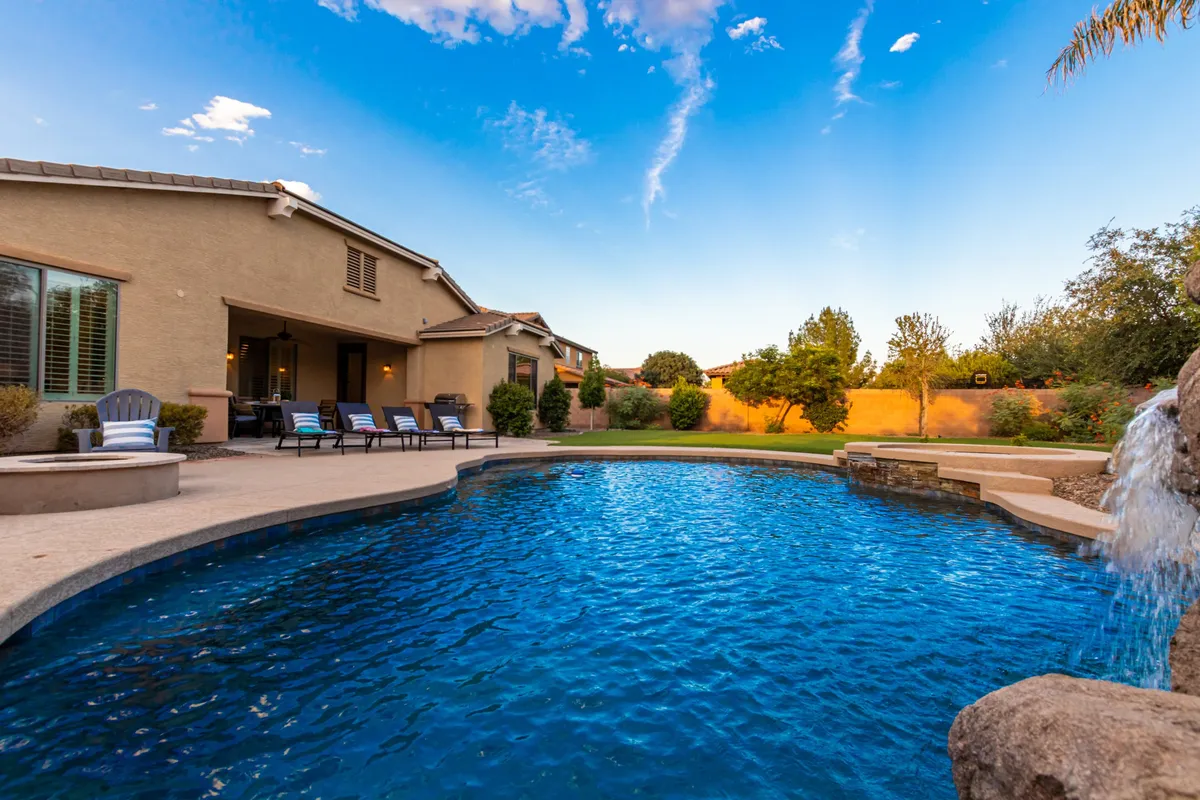 The exterior scene spotlights a sparkling pool with a waterfall feature, lounge chairs, and the home's façade. The landscaping and vibrant greenery add to the cheerful, resort-like ambiance, suggesting both daytime relaxation and lively evening barbecues.