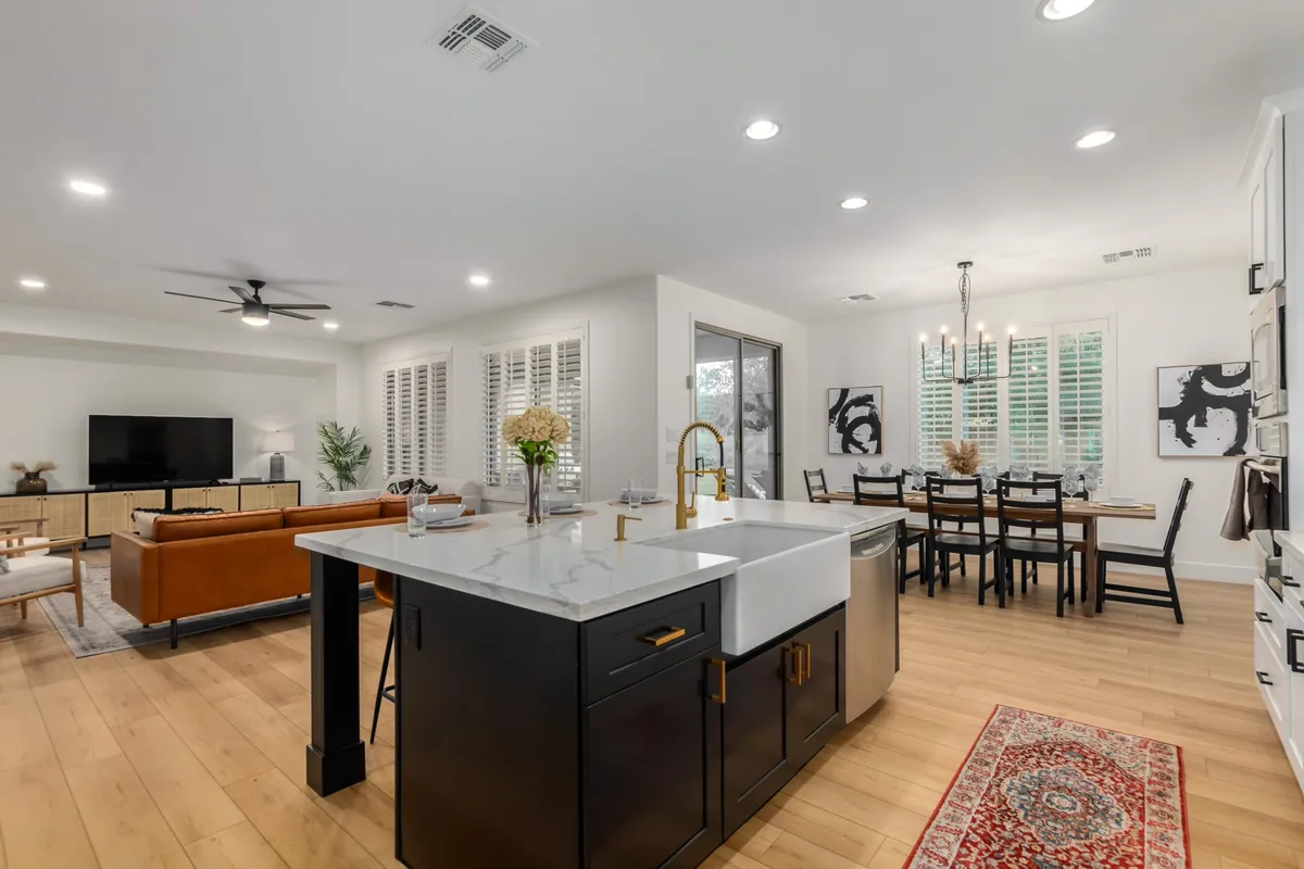 A wide, modern open-concept space that combines a kitchen island, dining table, and living room seating area. Large windows let in abundant natural light, and tasteful artwork adorns the walls, reinforcing the sense of togetherness and flow.