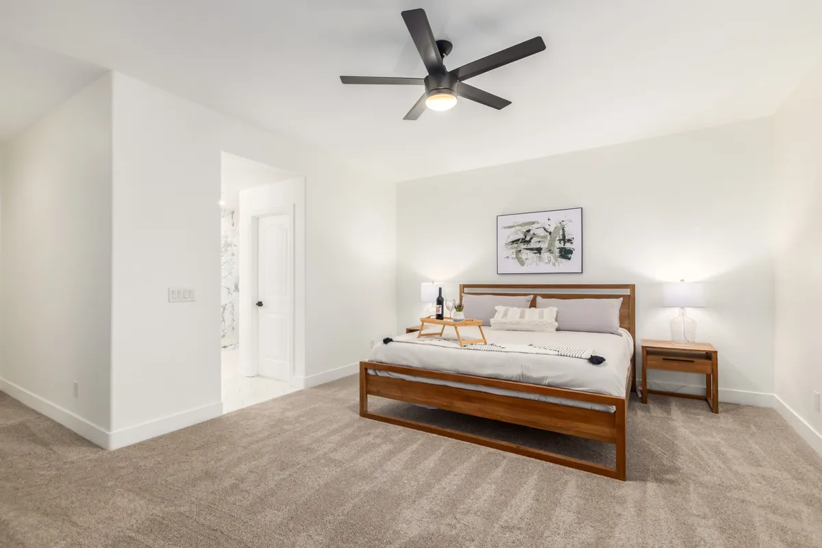 A tranquil bedroom featuring a plush bed flanked by nightstands, tasteful artwork above the headboard, and a ceiling fan. Sunlight streams through a window, creating a peaceful atmosphere ideal for restful nights.